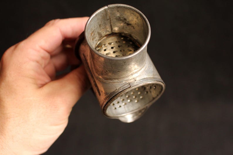 May include: Vintage metal kitchen tool with a cylindrical top and a perforated base. The tool is made of a silver-colored metal and has a T-shape. The tool is held in a hand against a dark background.