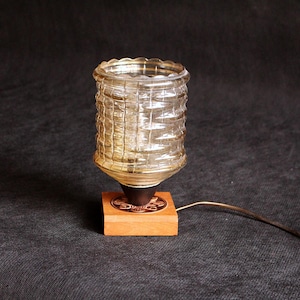May include: A vintage table lamp with a textured, cylindrical glass shade. The shade sits atop a dark brown base, mounted on a small, square wooden block. A cord with a white switch extends from the base.