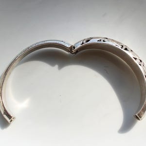 Puede incluir: Brazalete de plata articulado abierto con intrincados dise&ntilde;os florales y de volutas grabados. El brazalete se muestra sobre un fondo blanco, con el cierre abierto, revelando la superficie interior. El metal tiene un aspecto ligeramente envejecido.