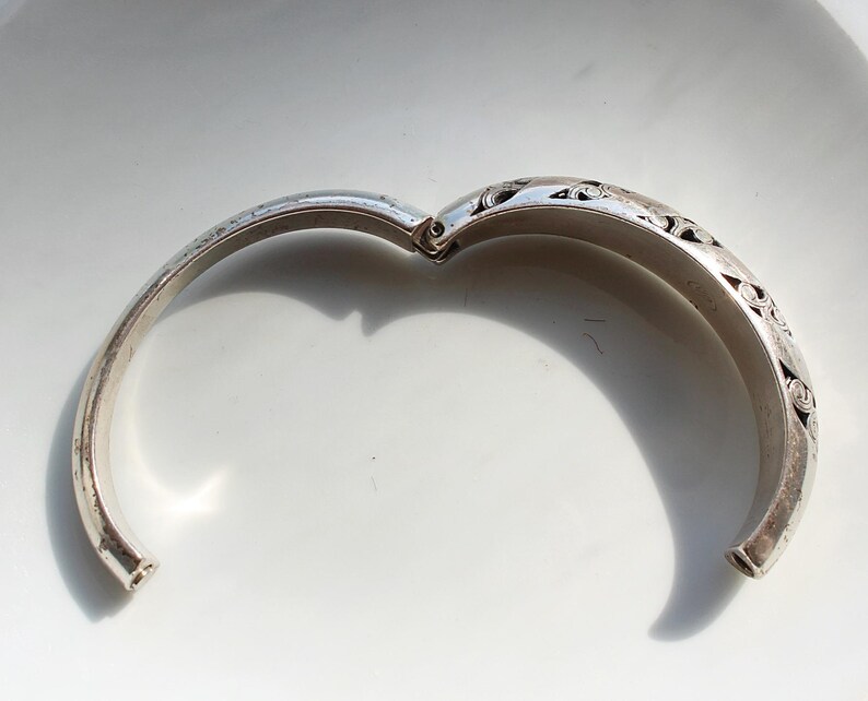 Puede incluir: Brazalete de plata articulado abierto con intrincados dise&ntilde;os florales y de volutas grabados. El brazalete se muestra sobre un fondo blanco, con el cierre abierto, revelando la superficie interior. El metal tiene un aspecto ligeramente envejecido.