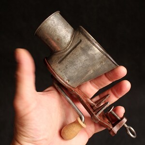 May include: An antique metal cheese grater with a wooden handle and clamp. The grater has a cylindrical top and a rectangular base with a crank. The metal has a worn, aged appearance.