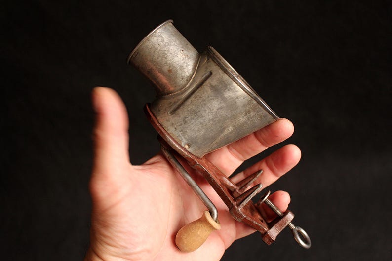 May include: An antique metal cheese grater with a wooden handle and clamp. The grater has a cylindrical top and a rectangular base with a crank. The metal has a worn, aged appearance.
