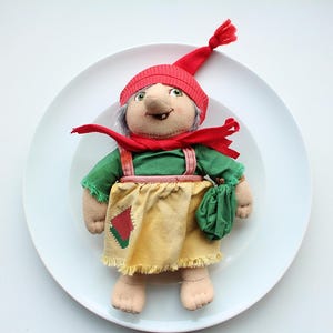 Puede incluir: Un gnomo de peluche con un gorro rojo, una camisa verde y una falda amarilla. El gnomo lleva una bolsa verde y está sentado en un plato blanco.