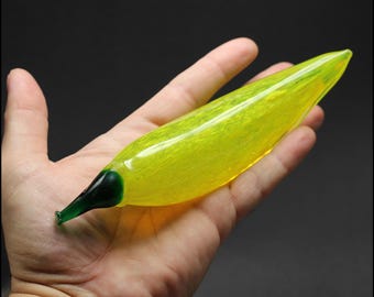 May include: A yellow glass banana with a green glass stem. The banana is slightly curved and has a textured surface.