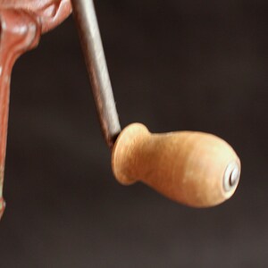 May include: Close-up of a vintage, cast iron hand crank with a wooden handle. The metal components are painted in a reddish-brown color, with a silver-colored crank shaft. The wooden handle is light brown.