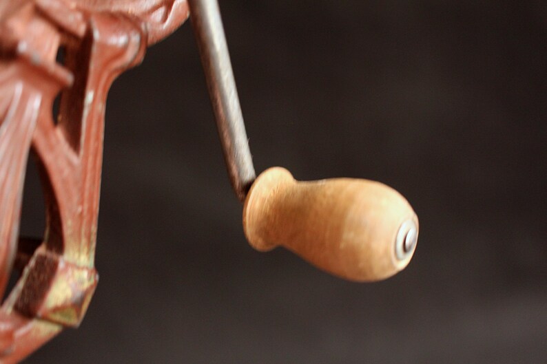 May include: Close-up of a vintage, cast iron hand crank with a wooden handle. The metal components are painted in a reddish-brown color, with a silver-colored crank shaft. The wooden handle is light brown.