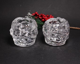 May include: Two clear, textured glass votive candle holders. The holders have a rough, organic shape and sit on a dark surface. Red berries and green sprigs are in the background. The candle holders are designed to hold small candles.