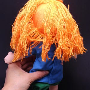Pode incluir: Boneca feita &agrave; m&atilde;o com cabelo de fio laranja, camisa azul, cal&ccedil;&otilde;es verdes e sapatos pretos. A boneca &eacute; segurada por uma m&atilde;o contra um fundo escuro. As caracter&iacute;sticas da boneca s&atilde;o simples e infantis.