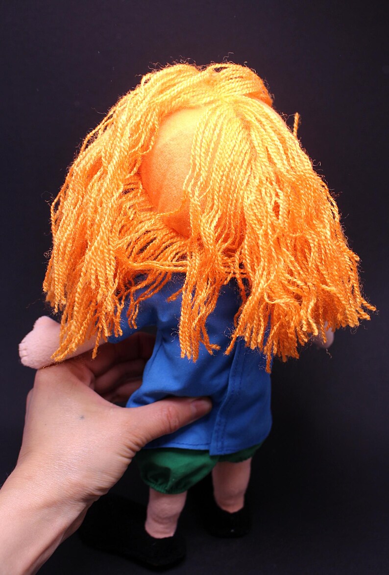 Pode incluir: Boneca feita &agrave; m&atilde;o com cabelo de fio laranja, camisa azul, cal&ccedil;&otilde;es verdes e sapatos pretos. A boneca &eacute; segurada por uma m&atilde;o contra um fundo escuro. As caracter&iacute;sticas da boneca s&atilde;o simples e infantis.