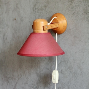 Könnte beinhalten: Eine Wandleuchte mit Holzsockel und rotem Lampenschirm. Die Lampe hat einen konischen Schirm und ein weißes Kabel mit einem Ein-/Ausschalter. Die Holzteile haben eine natürliche Oberfläche.