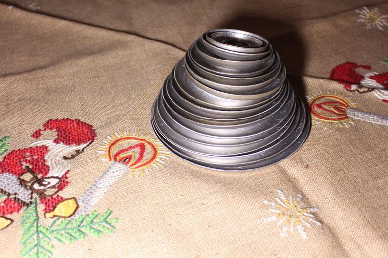 Puede incluir: Un conjunto de cortadores de galletas met&aacute;licos plateados graduados, dispuestos en forma c&oacute;nica. Los cortadores est&aacute;n sobre una tela beige con dise&ntilde;os navide&ntilde;os bordados, incluyendo una figura de Pap&aacute; Noel y copos de nieve.