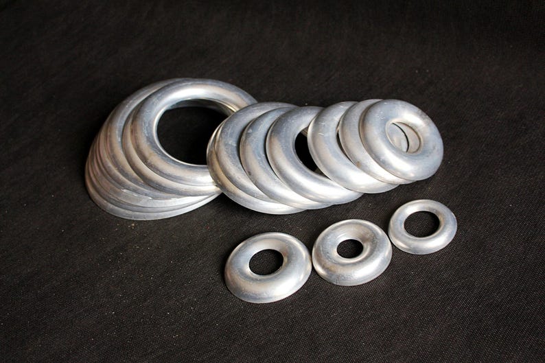 Puede incluir: Un conjunto de anillos met&aacute;licos plateados de varios tama&ntilde;os, dispuestos sobre una superficie negra. Los anillos tienen un dise&ntilde;o liso y redondeado, con un agujero en el centro. El conjunto de anillos m&aacute;s grande mide unos 10 cm de di&aacute;metro.