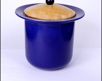 May include: A blue ceramic jar with a wooden lid and a black knob. The jar has a rounded shape and a wide mouth.