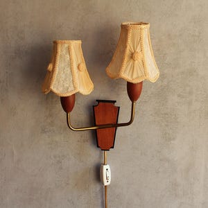 May include: Vintage wall sconce with two cream-colored lampshades. The shades have a scalloped edge and a floral accent. The sconce features a wooden backplate, brass arms, and a toggle switch. A classic lighting fixture.