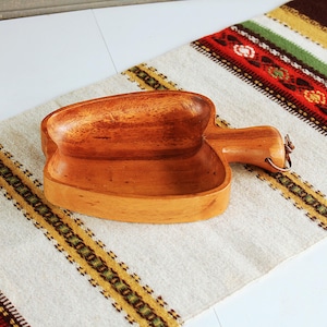 Op de afbeelding: Een houten kom in de vorm van een paprika met een handvat. De kom is gemaakt van lichtbruin hout en heeft een natuurlijke afwerking. De kom staat op een wit en bruin gestreept doek.