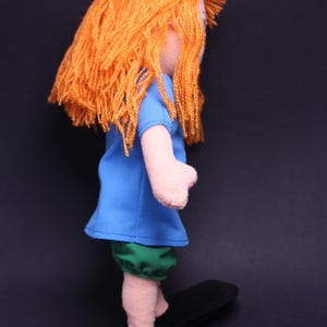 Pode incluir: Boneca feita &agrave; m&atilde;o com cabelo de fio laranja, vestindo uma blusa azul e shorts verdes. A boneca tem bra&ccedil;os e pernas rosa claro e sapatos pretos. A boneca est&aacute; em um fundo escuro.