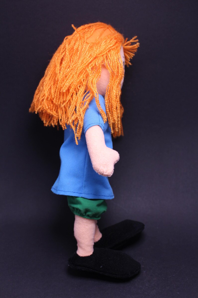 Pode incluir: Boneca feita &agrave; m&atilde;o com cabelo de fio laranja, vestindo uma blusa azul e shorts verdes. A boneca tem bra&ccedil;os e pernas rosa claro e sapatos pretos. A boneca est&aacute; em um fundo escuro.