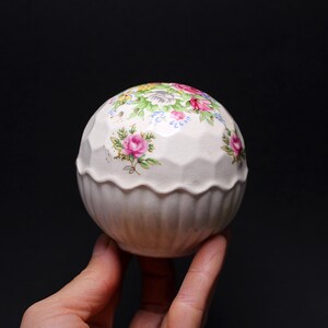 May include: A white, spherical salt shaker with a floral pattern featuring pink roses, blue flowers, and green leaves. The shaker has a textured, faceted design on the top half and a ribbed bottom. The salt shaker is held in a hand.