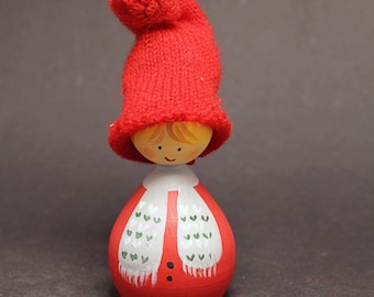 May include: A wooden doll with a red knitted hat and body. The doll features a painted face and a white scarf with green leaf patterns. A decorative item, it is approximately 15 cm tall.