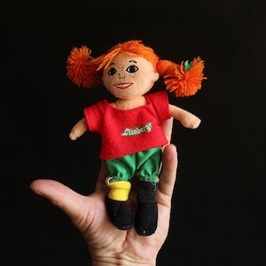 Può includere: Una piccola bambola con codini di filo arancione, che indossa una maglietta rossa con la scritta "Liseberg" ricamata, pantaloni verdi, calzini gialli e scarpe nere. La bambola è tenuta in mano su sfondo nero.