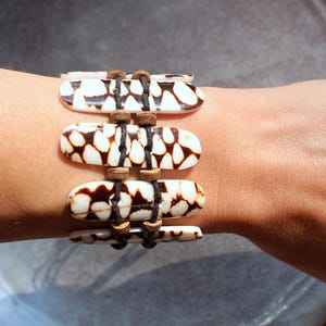 May include: A handcrafted bracelet featuring four oblong, cream-colored beads with brown patterned markings. The beads are connected by black cord and small wooden beads. This unique accessory is a stylish addition to any jewelry collection.