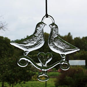 Puede incluir: Un adorno de vidrio con dos palomas posadas en una rama en forma de coraz&oacute;n con un dise&ntilde;o de ojo de cerradura. Las palomas se miran y parecen estar bes&aacute;ndose.
