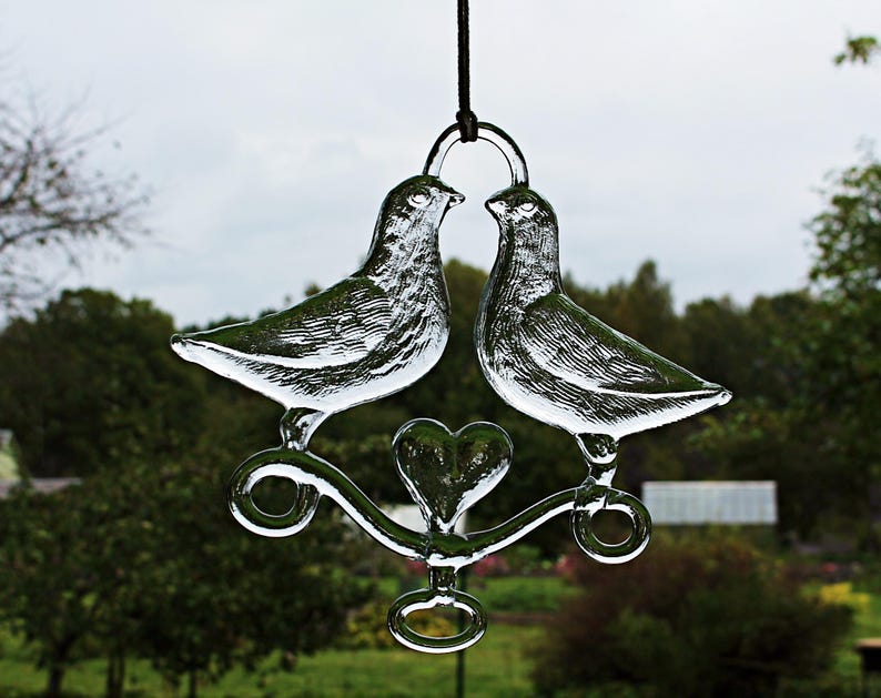 Puede incluir: Un adorno de vidrio con dos palomas posadas en una rama en forma de coraz&oacute;n con un dise&ntilde;o de ojo de cerradura. Las palomas se miran y parecen estar bes&aacute;ndose.