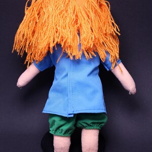 Pode incluir: Boneca feita &agrave; m&atilde;o com cabelo de fio laranja, uma blusa azul, cal&ccedil;&otilde;es verdes e sapatos pretos. Os bra&ccedil;os e as pernas da boneca s&atilde;o feitos de um tecido bege claro. A boneca est&aacute; em p&eacute; contra um fundo escuro.