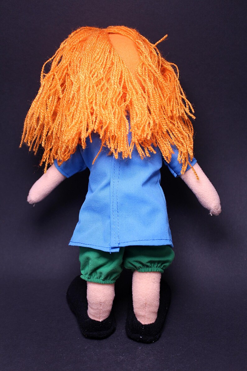 Pode incluir: Boneca feita &agrave; m&atilde;o com cabelo de fio laranja, uma blusa azul, cal&ccedil;&otilde;es verdes e sapatos pretos. Os bra&ccedil;os e as pernas da boneca s&atilde;o feitos de um tecido bege claro. A boneca est&aacute; em p&eacute; contra um fundo escuro.
