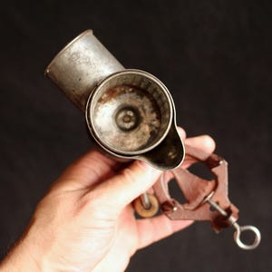 May include: An antique, hand-cranked cheese grater made of metal. The grater has a cylindrical body with a handle and a crank mechanism. The metal has a worn, aged appearance, suggesting its vintage. The grater is held against a dark background.