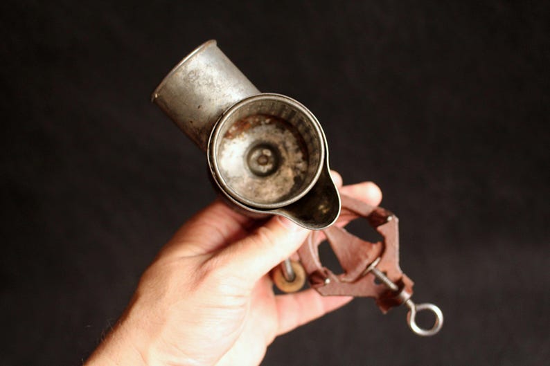 May include: An antique, hand-cranked cheese grater made of metal. The grater has a cylindrical body with a handle and a crank mechanism. The metal has a worn, aged appearance, suggesting its vintage. The grater is held against a dark background.