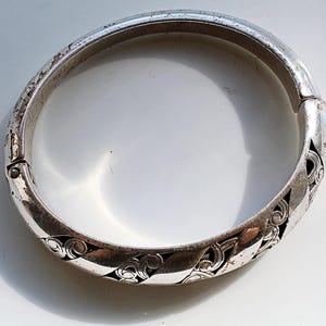 Puede incluir: Una pulsera de brazalete plateada con bisagras y un dise&ntilde;o calado ornamentado. La pulsera presenta un patr&oacute;n repetitivo de formas arremolinadas en espiral. El metal tiene un aspecto ligeramente envejecido, con algo de desgaste visible en la superficie.