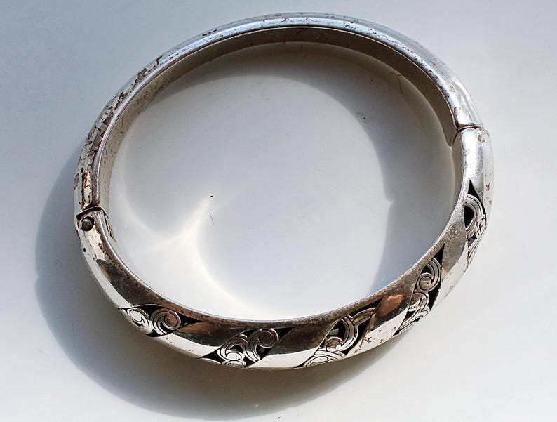 Puede incluir: Una pulsera de brazalete plateada con bisagras y un dise&ntilde;o calado ornamentado. La pulsera presenta un patr&oacute;n repetitivo de formas arremolinadas en espiral. El metal tiene un aspecto ligeramente envejecido, con algo de desgaste visible en la superficie.