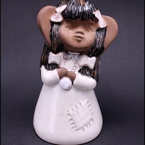 Puede incluir: Una figurita de cerámica de una niña con piel morena, cabello negro y un vestido blanco. Lleva un lazo blanco a cada lado de la cabeza y tiene las manos juntas frente a ella. El vestido tiene un parche en forma de corazón en la parte delantera.