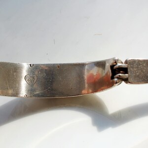 Puede incluir: Una pulsera articulada plateada con un emblema en forma de coraz&oacute;n. La pulsera es curva y tiene un cierre rectangular. El metal tiene un aspecto ligeramente envejecido, con algunas imperfecciones menores visibles.