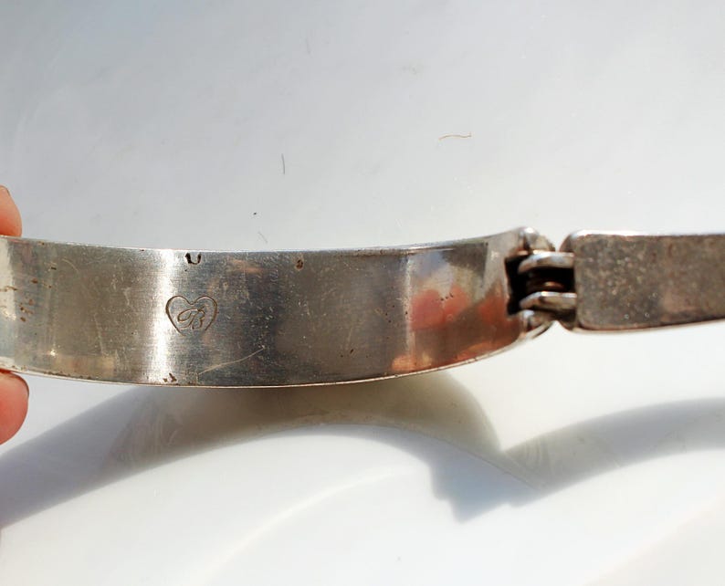 Puede incluir: Una pulsera articulada plateada con un emblema en forma de coraz&oacute;n. La pulsera es curva y tiene un cierre rectangular. El metal tiene un aspecto ligeramente envejecido, con algunas imperfecciones menores visibles.