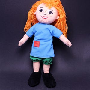 May include: A handmade doll with orange yarn hair, a blue shirt with a red square patch, green shorts, and black shoes. The doll has a painted face with freckles and a small smile. A soft toy for children.