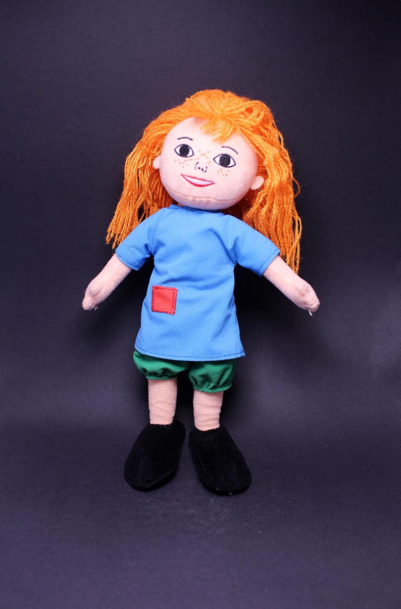 Pode incluir: Boneca feita &agrave; m&atilde;o com cabelo de fio laranja, uma camisa azul com um remendo quadrado vermelho, cal&ccedil;&otilde;es verdes e sapatos pretos. A boneca tem um rosto pintado com sardas e um pequeno sorriso. Um brinquedo macio para crian&ccedil;as.