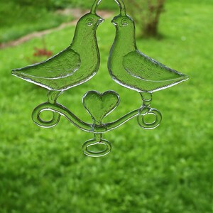 Puede incluir: Dos palomas de vidrio transparente sujetando un amuleto en forma de coraz&oacute;n, colgando de una cuerda. Las palomas se miran y sus picos se tocan. El amuleto en forma de coraz&oacute;n est&aacute; en el centro de las palomas.