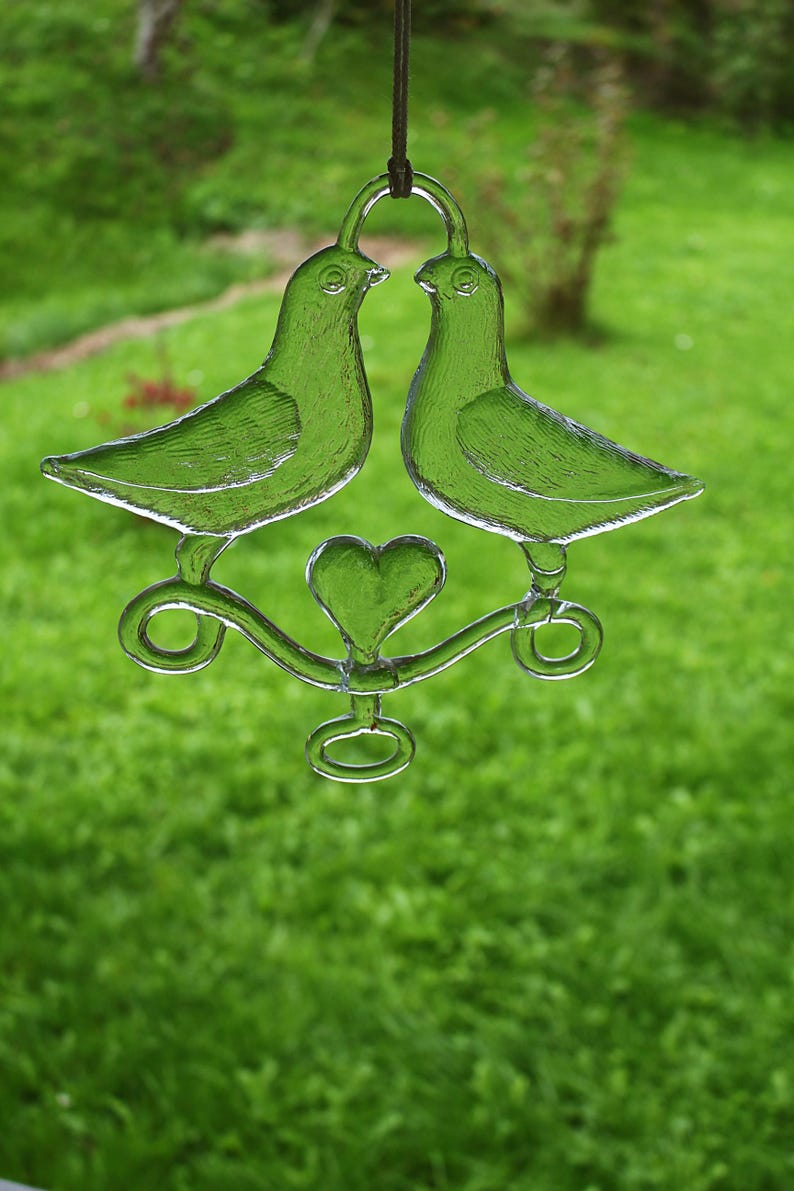 Puede incluir: Dos palomas de vidrio transparente sujetando un amuleto en forma de coraz&oacute;n, colgando de una cuerda. Las palomas se miran y sus picos se tocan. El amuleto en forma de coraz&oacute;n est&aacute; en el centro de las palomas.