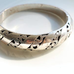 Puede incluir: Un brazalete de bisagra plateado. El brazalete presenta un patr&oacute;n repetitivo de dise&ntilde;os calados estilizados con un fondo oscuro. El brazalete es circular y parece estar hecho de metal.