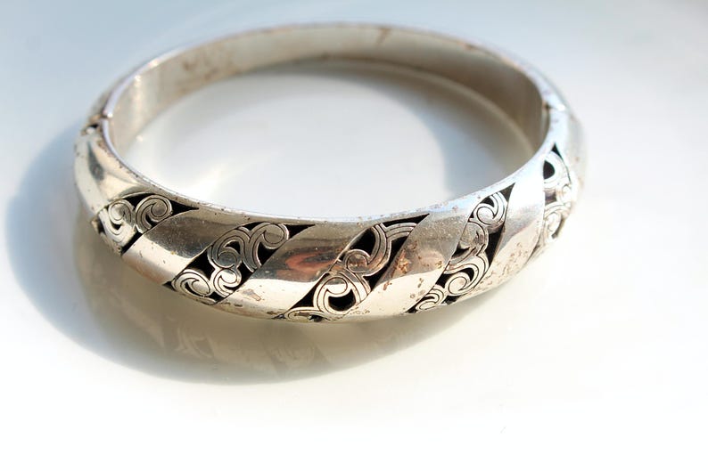 Puede incluir: Un brazalete de bisagra plateado. El brazalete presenta un patr&oacute;n repetitivo de dise&ntilde;os calados estilizados con un fondo oscuro. El brazalete es circular y parece estar hecho de metal.