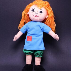 Pode incluir: Boneca feita &agrave; m&atilde;o com cabelo de fio laranja, um rosto sorridente e sardas. A boneca usa uma t&uacute;nica azul com um remendo quadrado vermelho, shorts verdes e sapatos pretos. Os bra&ccedil;os da boneca est&atilde;o estendidos.