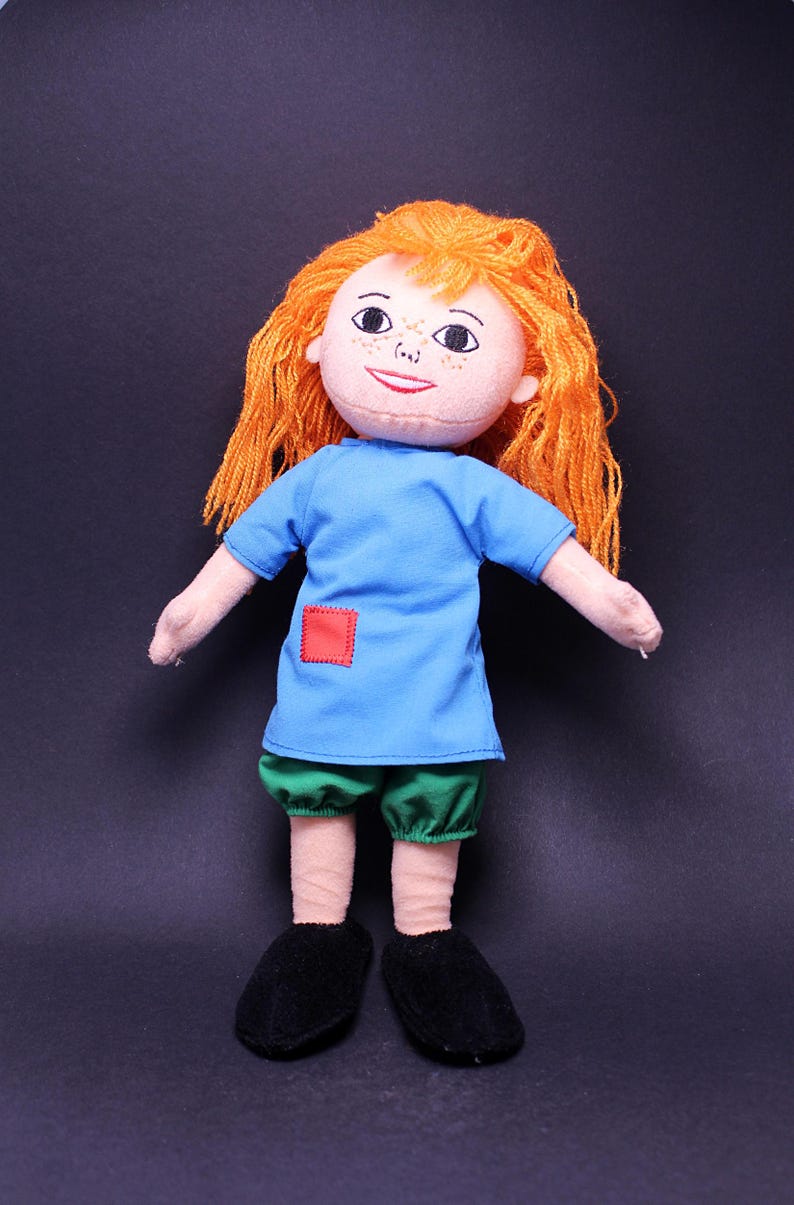 Pode incluir: Boneca feita &agrave; m&atilde;o com cabelo de fio laranja, um rosto sorridente e sardas. A boneca usa uma t&uacute;nica azul com um remendo quadrado vermelho, shorts verdes e sapatos pretos. Os bra&ccedil;os da boneca est&atilde;o estendidos.