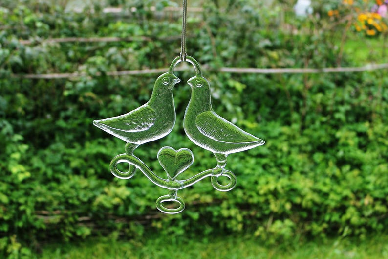 Puede incluir: Un adorno de vidrio con dos palomas y un coraz&oacute;n en el centro. Las palomas son de vidrio transparente y el coraz&oacute;n es esmerilado.