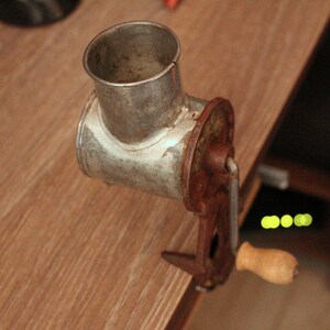 May include: An antique metal meat grinder with a rusty brown handle and a wooden grip. The grinder has a cylindrical opening and a clamp to secure it to a surface. The metal has a worn, aged appearance.