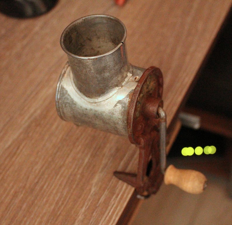May include: An antique metal meat grinder with a rusty brown handle and a wooden grip. The grinder has a cylindrical opening and a clamp to secure it to a surface. The metal has a worn, aged appearance.
