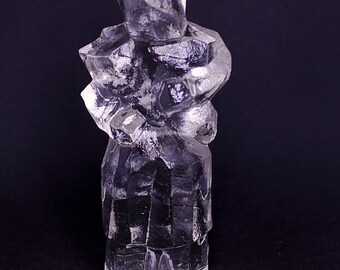May include: A clear, faceted glass sculpture of a person. The figure is depicted in a blocky, geometric style, with visible edges and angles. The sculpture is set against a dark background, highlighting its transparent form.