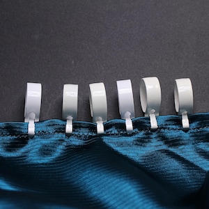 May include: White curtain rings attached to a teal fabric. The rings are evenly spaced and appear to be made of metal. The fabric has a shiny, silky texture. The background is a dark gray color.