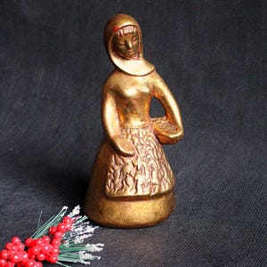 Può includere: Statuetta color oro di una donna con copricapo e grembiule, che tiene un cestino. La figura ha un design dettagliato del grembiule ed è ambientata su uno sfondo scuro. Un rametto di bacche rosse e accenti bianchi è in primo piano.